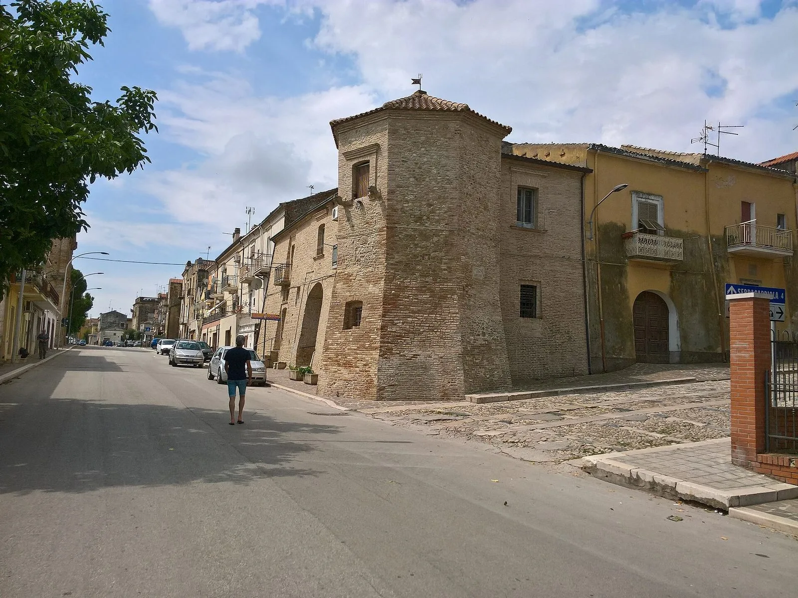 Una strada di Chieuti con un'antica torre in mattoni e pietra dal tetto in coppi, case gialle con balconi e un cartello stradale che indica Serracapriola