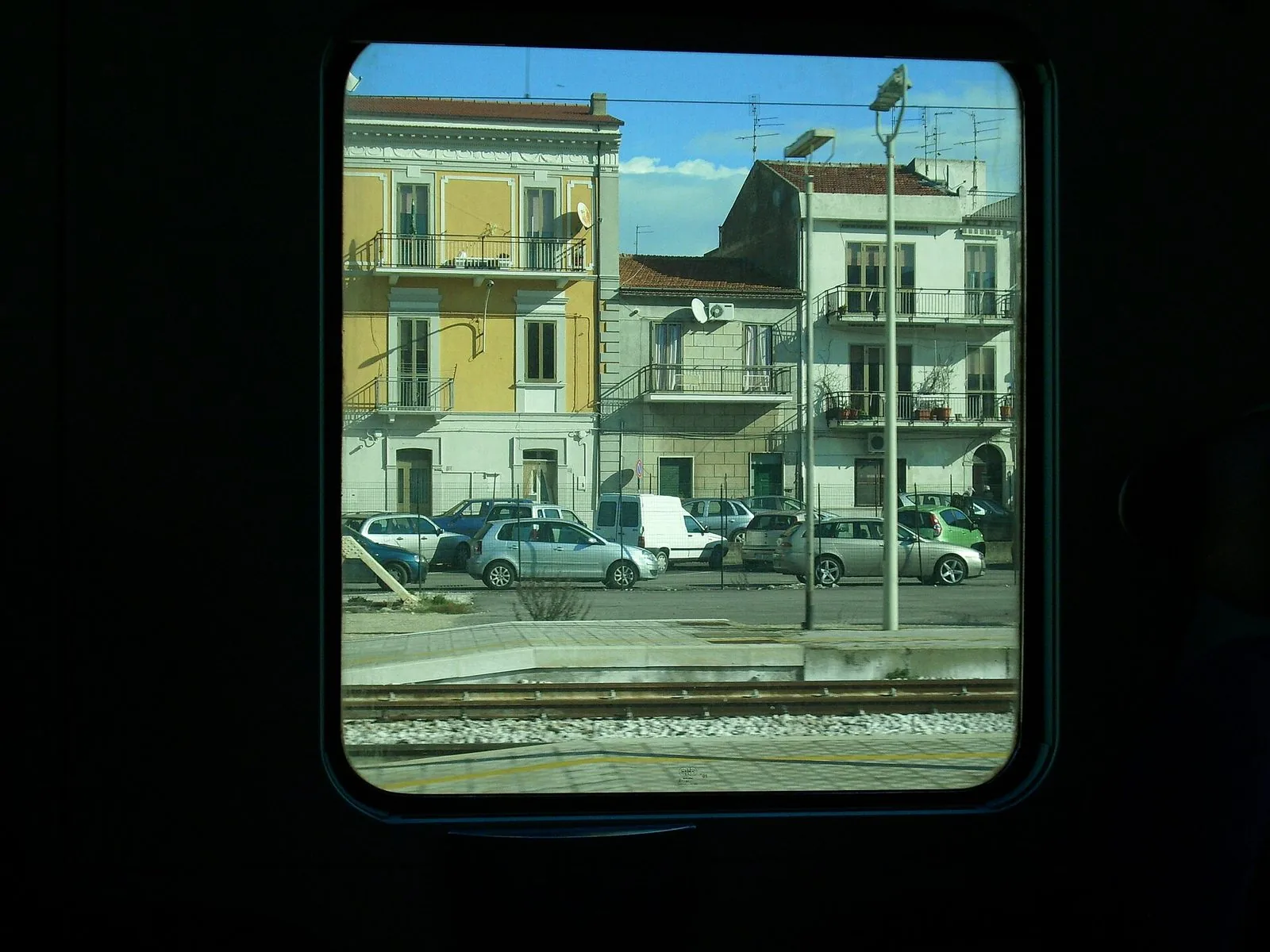Vista dal finestrino di un treno fermo alla stazione di Termoli: binari, marciapiede e palazzine colorate con auto parcheggiate