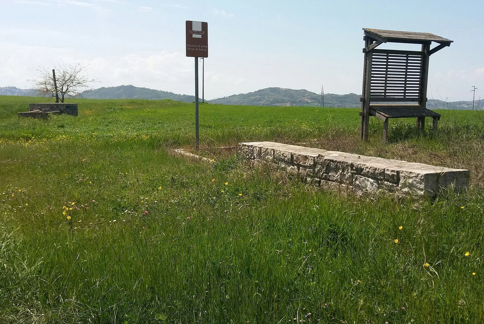 Un tratto erboso di regio tratturo con un abbeveratoio in pietra, una pensilina di legno e il cartello marrone di una piazzola di sosta, sullo sfondo colline verdi